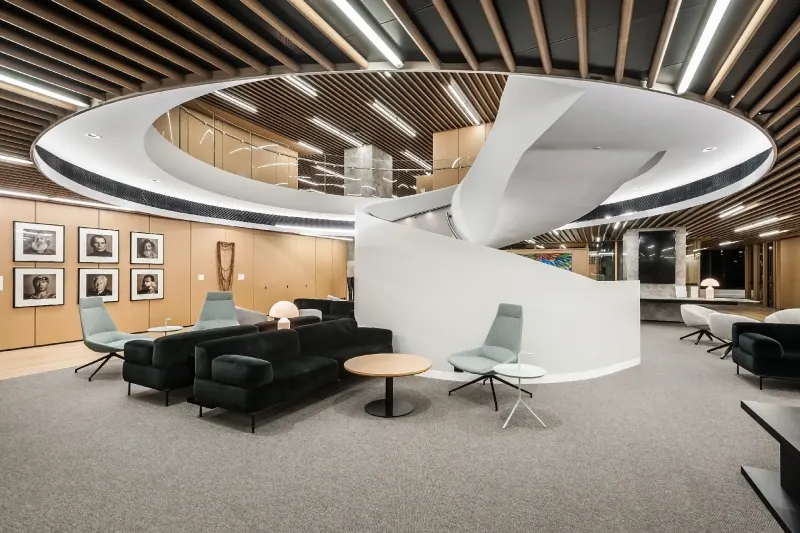 Workplace breakout lounge with dark sofas beneath a circular timber ceiling and spiral stair